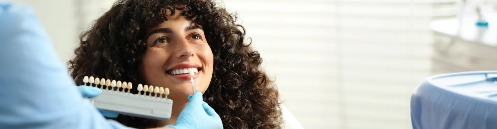 Gloved hand holding tooth shade-matching tool against woman's smile.