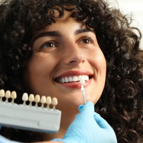Gloved hand holding tooth shade-matching tool against woman's smile.