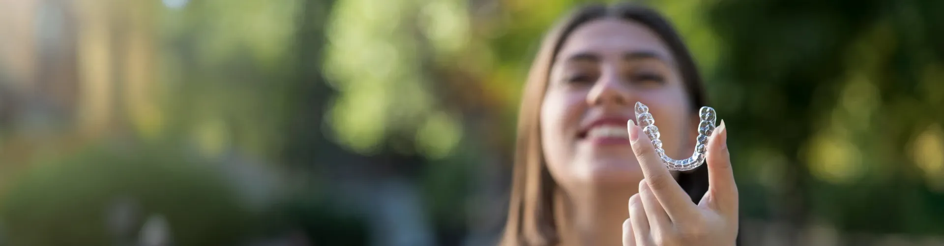Smiling woman outdoors holding a clear Invisalign aligner toward the camera, highlighting discreet orthodontic treatment.