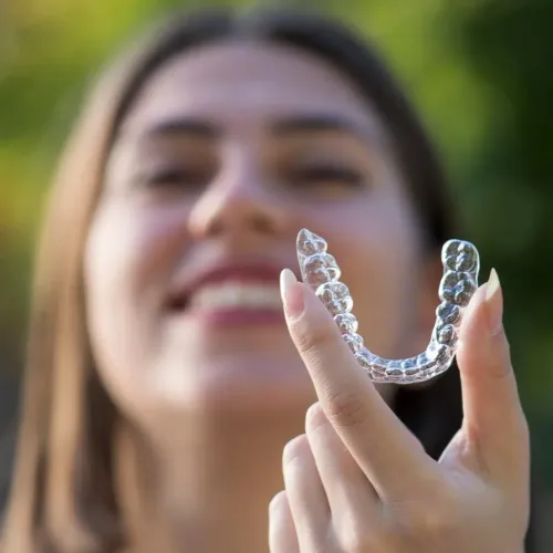 Smiling woman outdoors holding a clear Invisalign aligner toward the camera, highlighting discreet orthodontic treatment.
